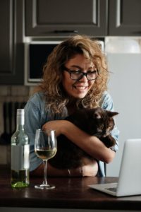 A person holding a cat on her lap whilst looking at a laptop, drinking a glass of wine during the video call.